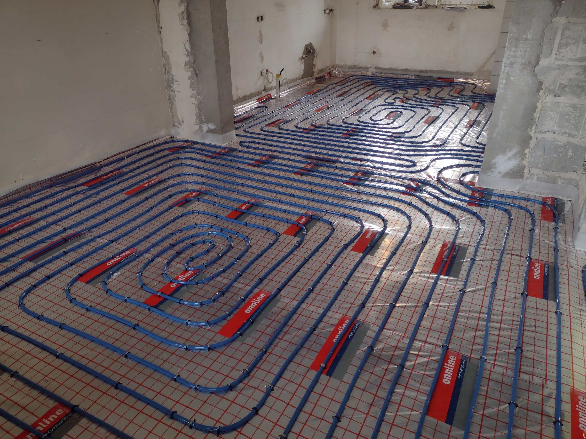 Niedokończona instalacja ogrzewania podłogowego z rurami w kolorze niebieskim, zamocowanymi na siatce z czerwonymi elementami mocującymi z logo 'onnline Professional', w surowym wnętrzu przed wylewką.