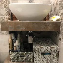 Nowoczesna umywalka nablatowa w kształcie misy, zamontowana na blacie z płytek imitujących kamień, dekoracyjne płytki ścienne w odcieniach szarości, widoczne akcesoria łazienkowe w ozdobnych...