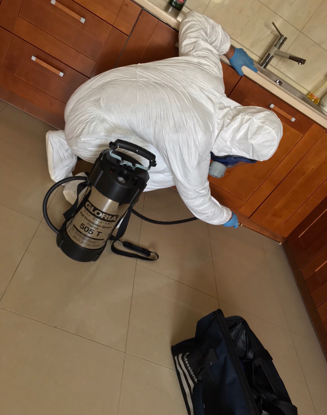 Pracownik w kombinezonie ochronnym i masce, z niebieskimi rękawicami, aplikuje preparat za pomocą dyszy przy listwie przypodłogowej szafki kuchennej; obok stoi opryskiwacz ciśnieniowy Gloria 505 T...