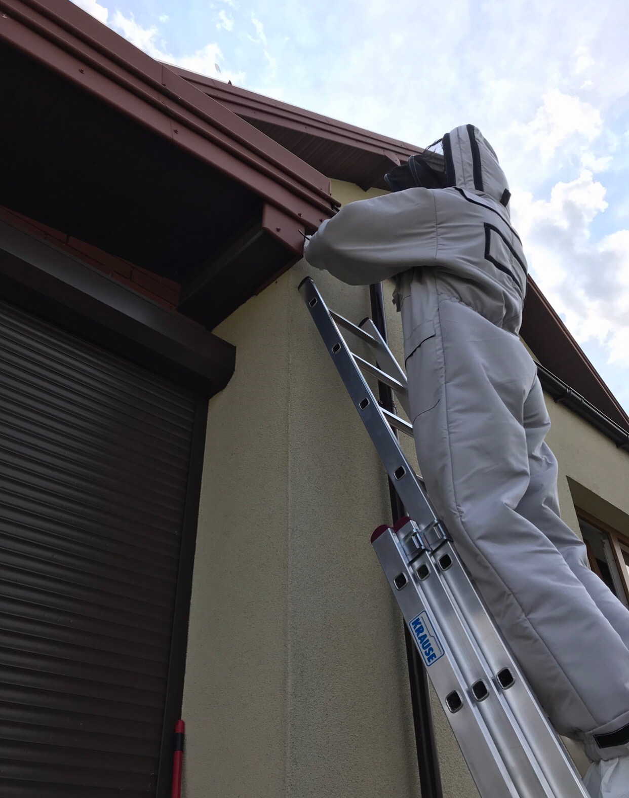 Pracownik w kombinezonie ochronnym na aluminiowej drabinie marki Krause, uszczelnia okap dachu przy użyciu narzędzia, widoczne brązowe rolety i fragment muru budynku.