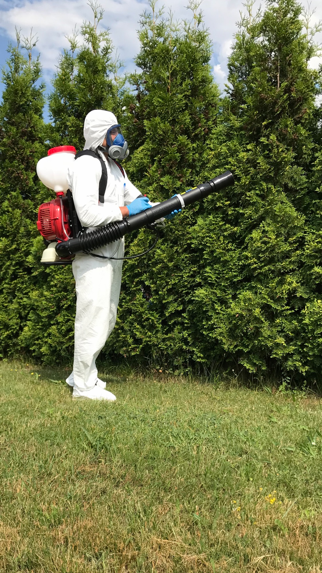 Osoba w kombinezonie ochronnym i masce, z opryskiwaczem plecakowym, aplikuje środek na zielone krzewy w ogrodzie.