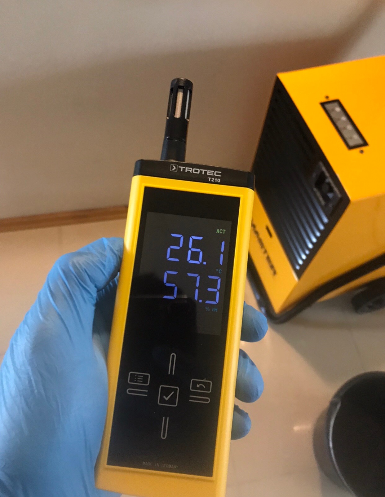 Miernik wilgotności Trotec T210 w dłoni w niebieskiej rękawiczce, wskazujący temperaturę 26.1°C i wilgotność 57.3% rH, z żółtym osuszaczem powietrza w tle.