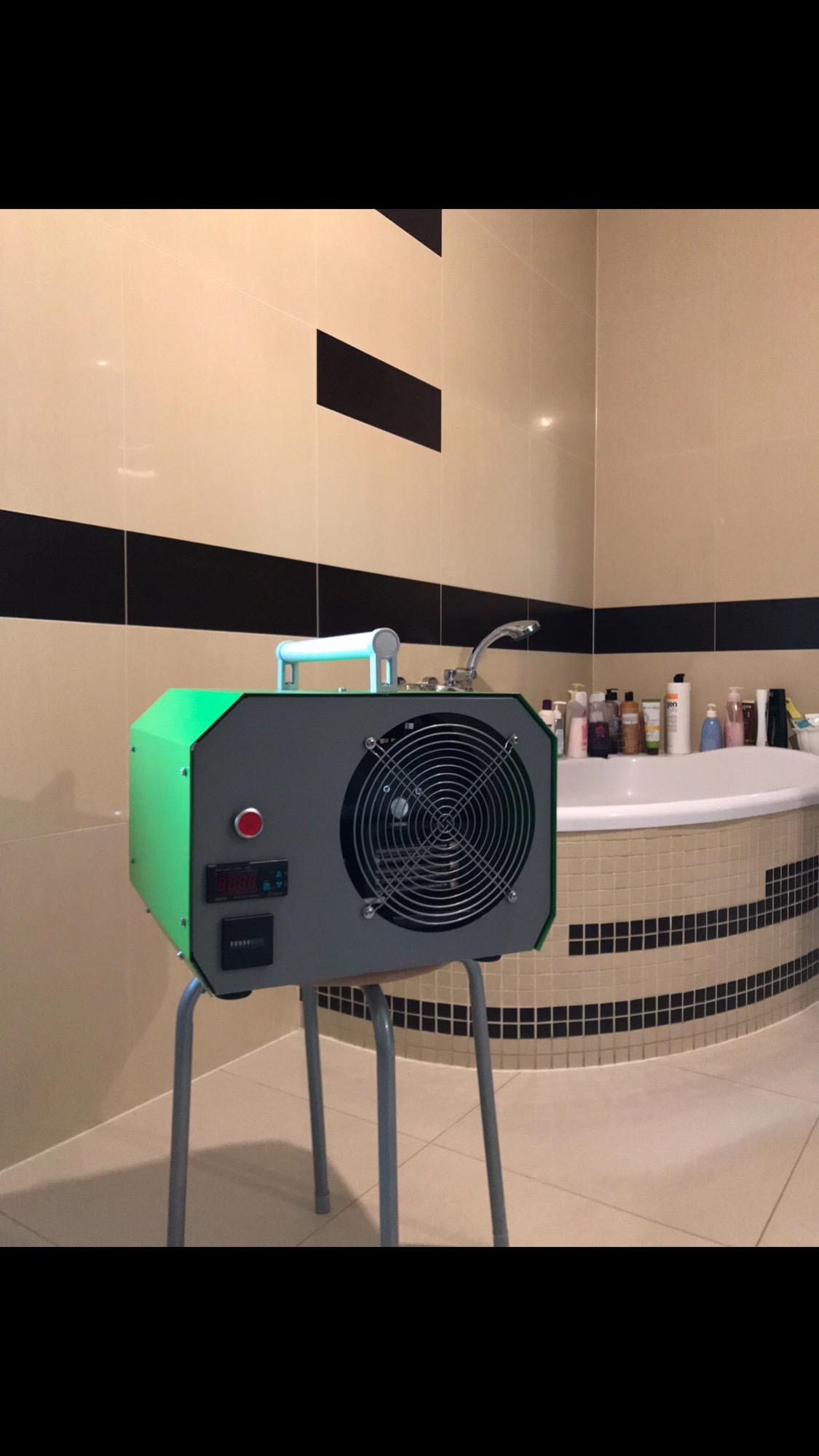 Profesjonalny osuszacz powietrza z zielonymi elementami obudowy, umieszczony na metalowym stołku w łazience z beżowo-czarnymi płytkami, obok wanny wypełnionej kosmetykami.