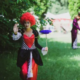 Osoba w kostiumie Królowej Kier z czerwonym afro i makijażem, trzymająca talerz na kiju, na tle zieleni i namiotów piknikowych, w tle postać w fioletowym fraku.