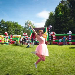 Kobieta w różowej sukience skacze na tle dmuchanych zamków w kształcie słodyczy podczas pikniku firmowego.