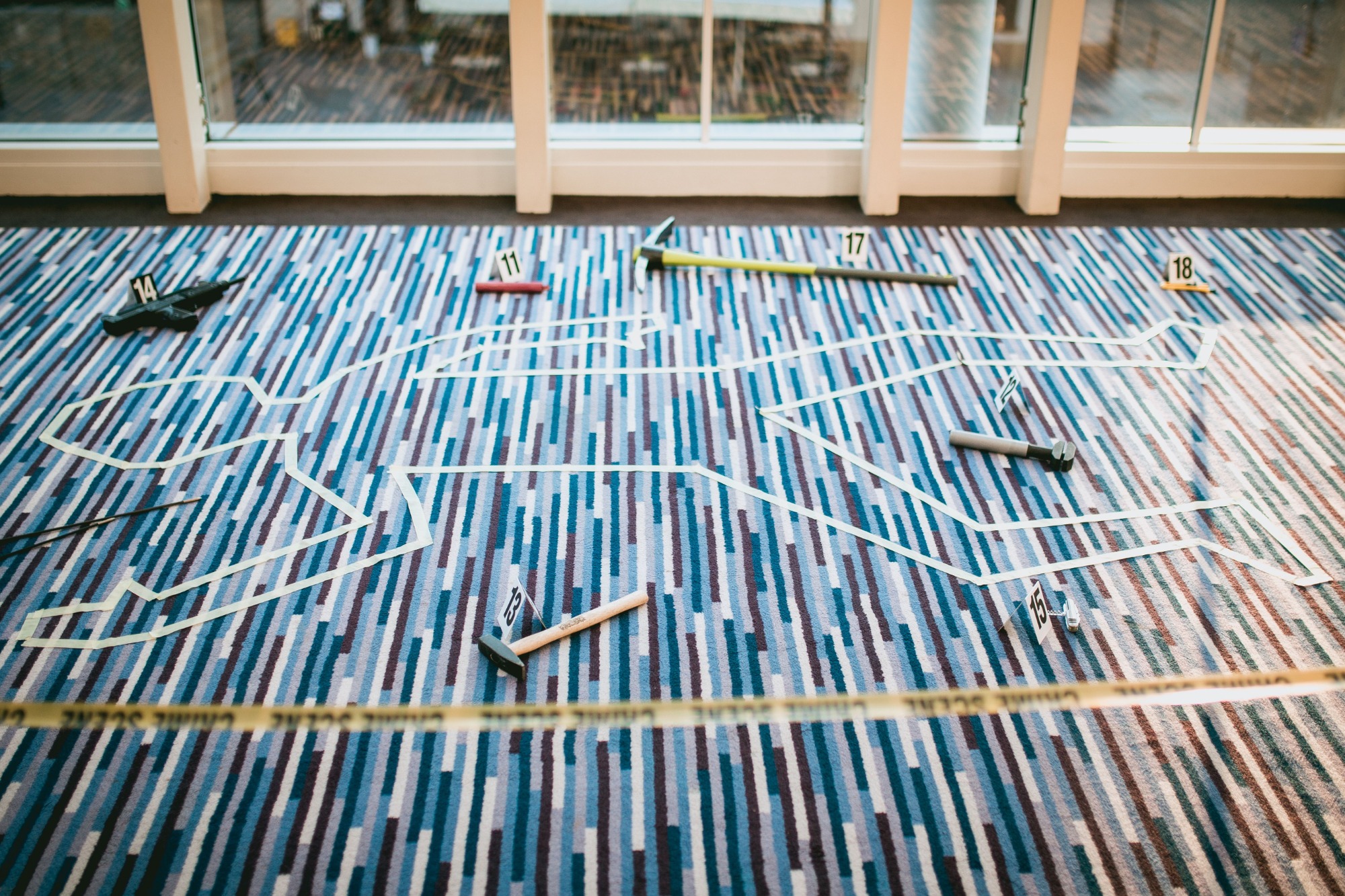 Ułożona na wzorzystym dywanie scena z gry szpiegowskiej, z obrysami ciał, rekwizytami oznaczonymi numerami i taśmą ostrzegawczą.