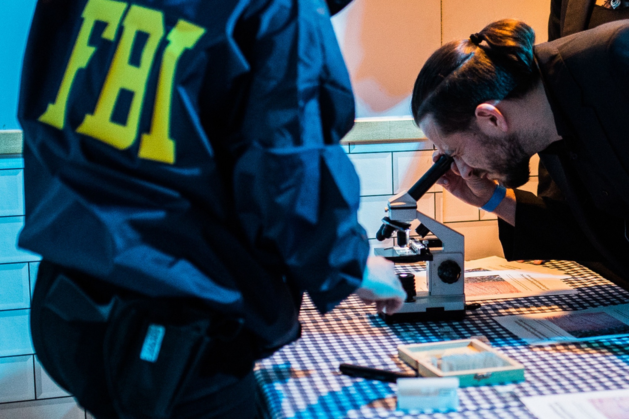 Osoba w kurtce z napisem FBI obserwuje próbkę pod mikroskopem na stole w niebiesko-białą kratę, druga osoba w garniturze również patrzy w mikroskop.