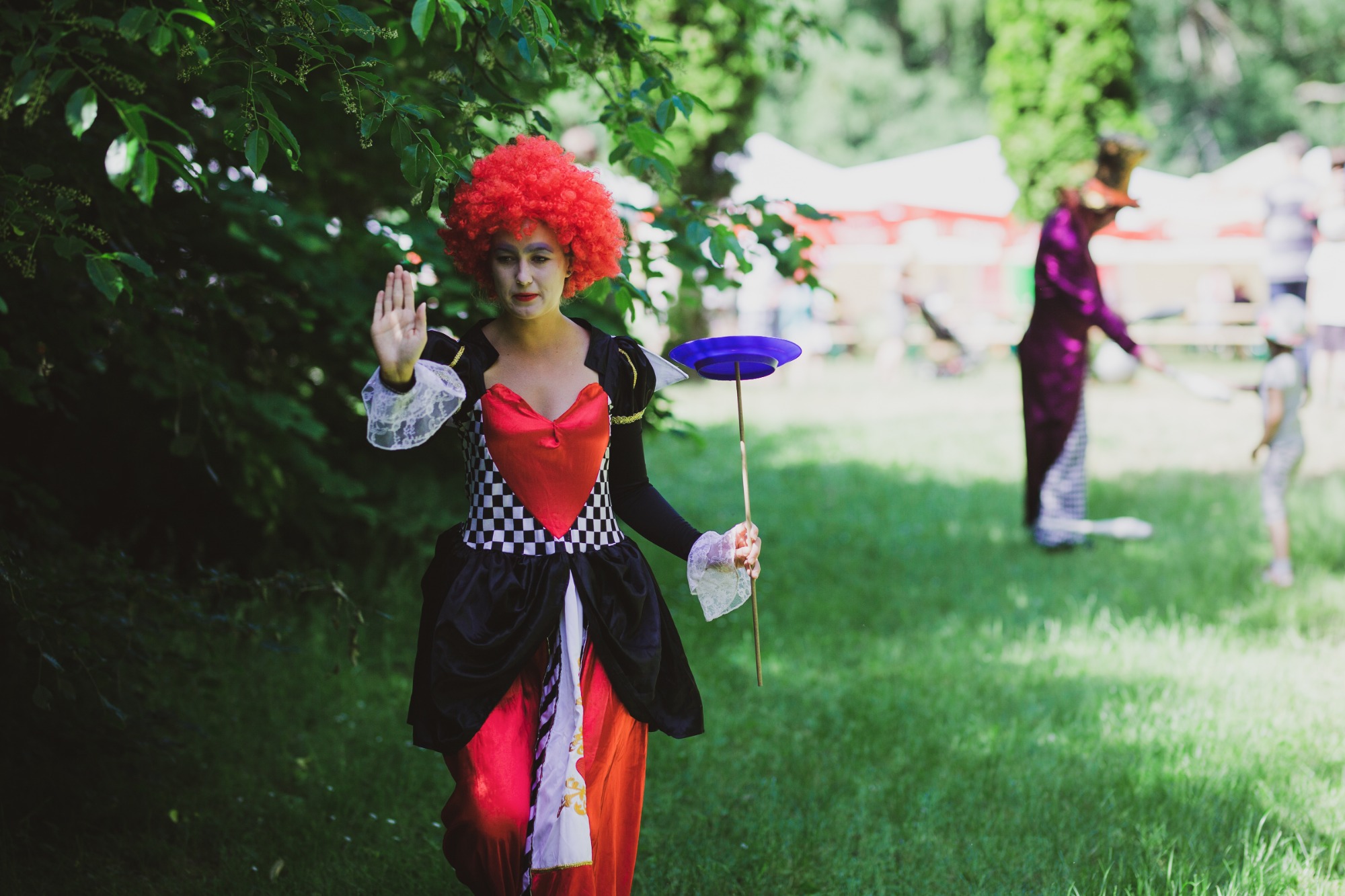 Osoba w kostiumie Królowej Kier z czerwonym afro i makijażem, trzymająca talerz na kiju, na tle zieleni i namiotów piknikowych, w tle postać w fioletowym fraku.