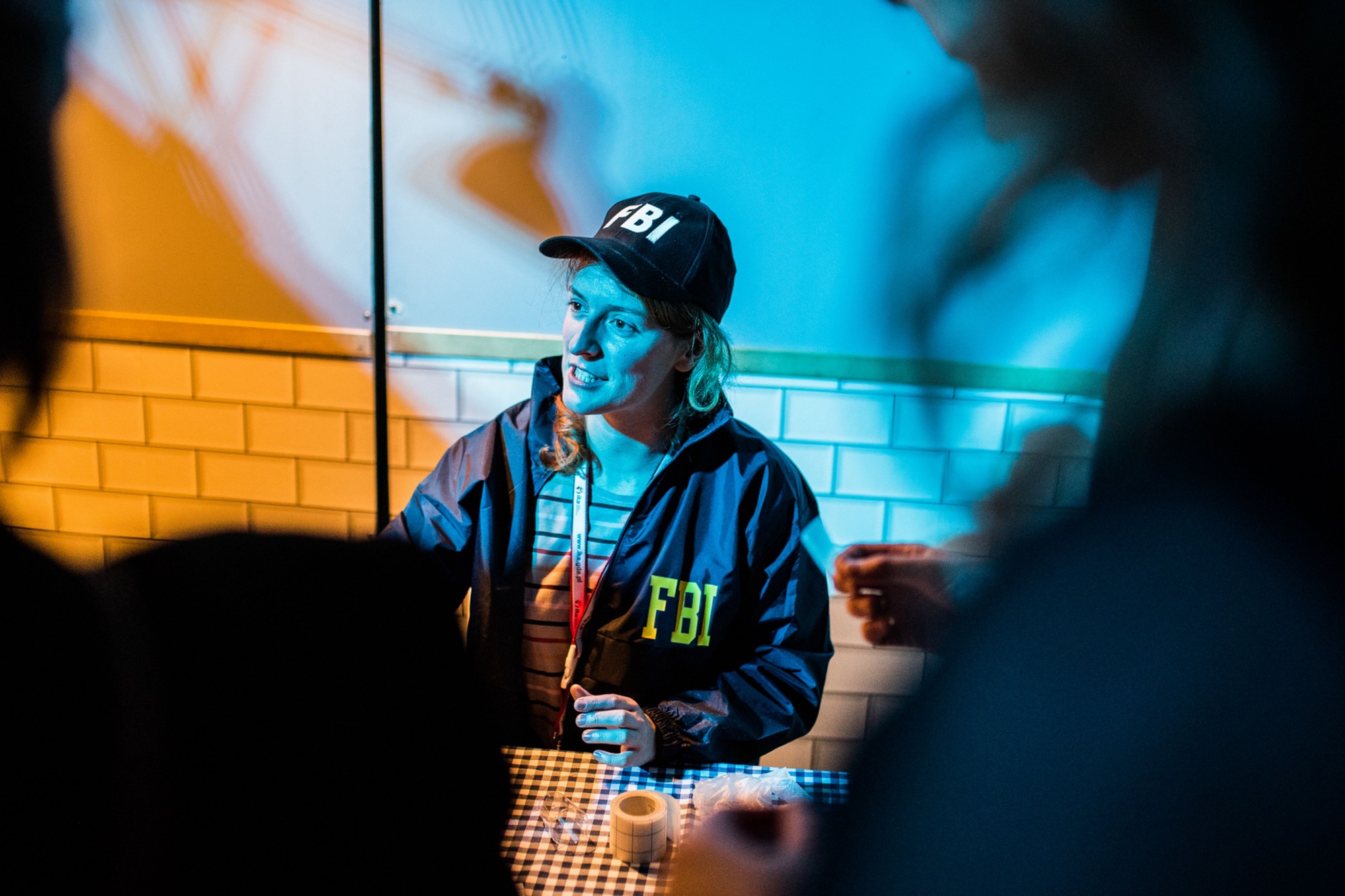 Osoba w czapce i kurtce z napisem FBI siedzi przy stole w kratę z taśmą klejącą, oświetlona niebieskim światłem, w tle białe kafelki.