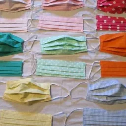 Różnokolorowe, bawełniane maseczki ochronne, ułożone rzędami na jasnej powierzchni, widoczne gumki mocujące.