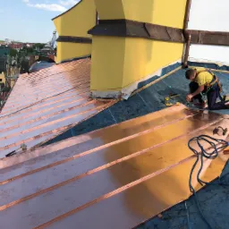 Dekarz w uprzęży montuje miedziane blachy na dachu budynku z żółtymi kominami, widok na miasto w tle.