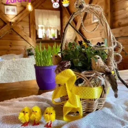 Świąteczny koszyk z dekoracjami: pisanki, ptaszki, żółta wstążka w kropki, trawa w fioletowej doniczce, trzy małe żółte kurczaczki na białym obrusie z koronką, drewniane wnętrze w tle.