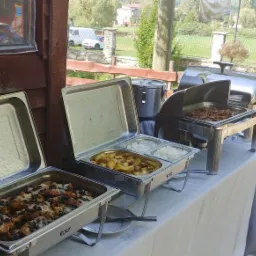 Bufet z różnorodnymi potrawami w metalowych bemarach: zapiekanka z warzywami, ziemniaki pieczone, ryż oraz danie mięsne, ustawiony na białym obrusie na zewnątrz.