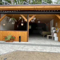 Zadaszona przestrzeń tarasowa z drewnianą konstrukcją, wyposażona w zwiniętą, czarną roletę, dekoracje w postaci lampionów i wiszących roślin, z widoczną kostką brukową na podłożu.