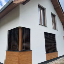 Nowoczesny dom jednorodzinny z białymi ścianami i brązowymi żaluzjami fasadowymi, widoczne niedokończone prace wykończeniowe wokół budynku.