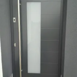 Nowoczesne, antracytowe drzwi zewnętrzne z długim, pionowym uchwytem ze stali nierdzewnej i prostokątnym przeszkleniem z matowego szkła.