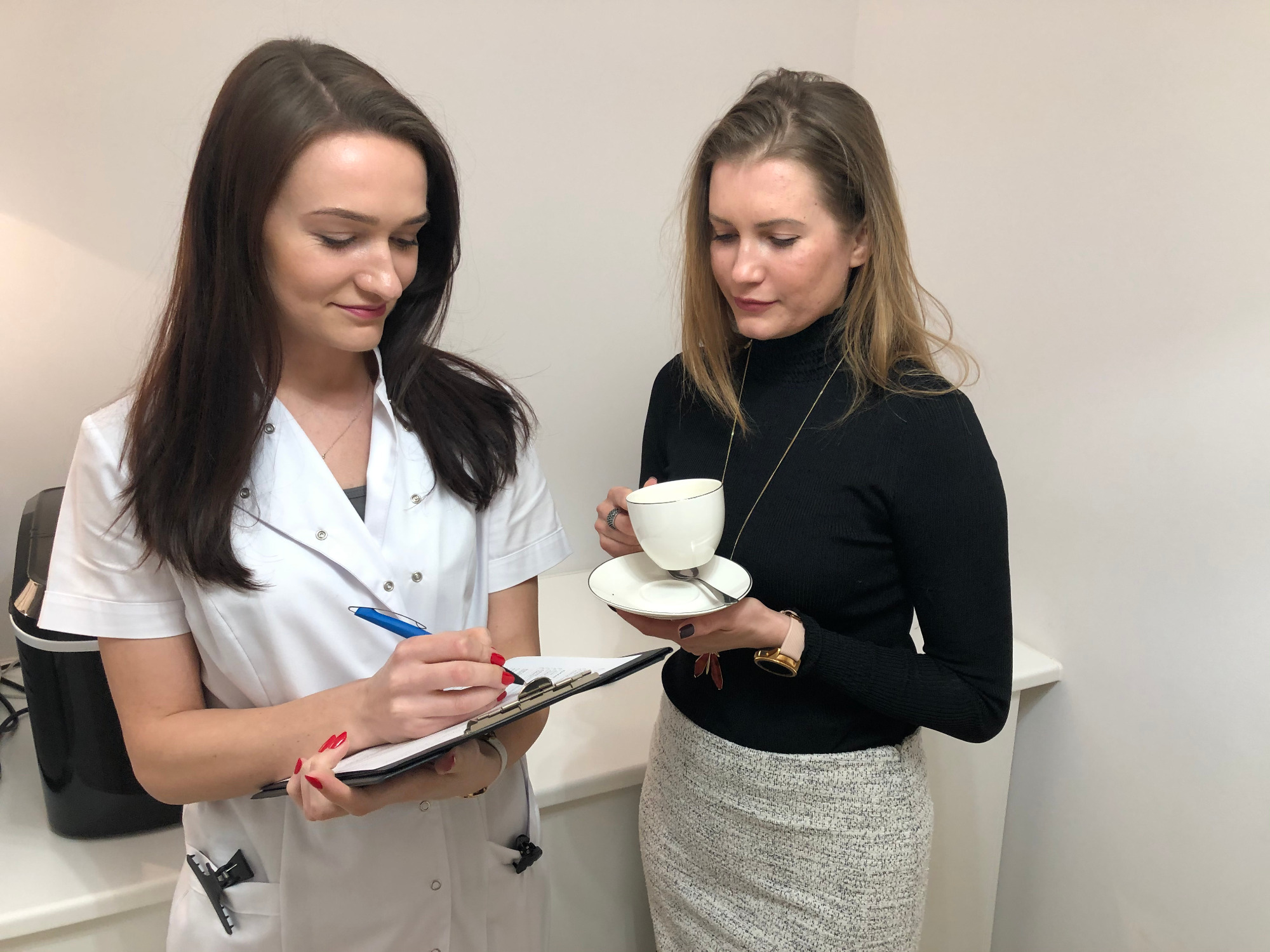 Konsultacja w gabinecie medycyny estetycznej: Lekarz w białym uniformie z długopisem i clipboardem omawia plan zabiegu z pacjentką trzymającą filiżankę kawy.