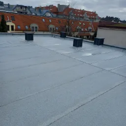 Szary, płaski dach pokryty papą z czterema ciemnymi kominkami wentylacyjnymi na tle budynków z czerwonymi dachami i pochmurnego nieba.