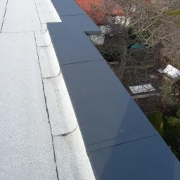 Szary metalowy element wykończeniowy na płaskim dachu pokrytym jasnym, szorstkim materiałem, z widokiem na drzewo bez liści i dach sąsiedniego budynku w tle.