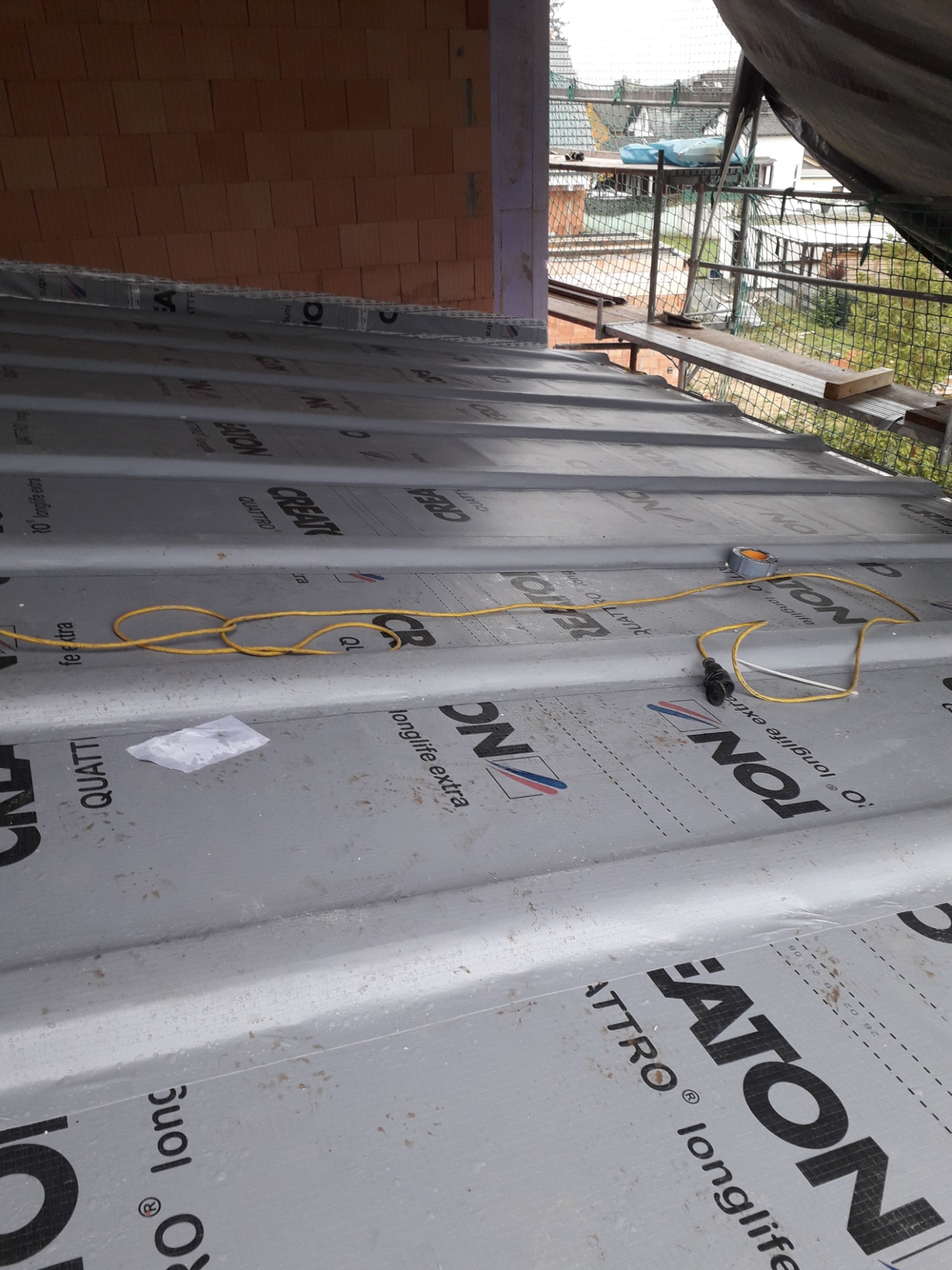 Widok z góry na przygotowaną podłoże pod pokrycie dachu, widoczne membrany dachowe z napisami, przewód elektryczny i taśma klejąca. W tle elementy konstrukcji budynku i rusztowania.