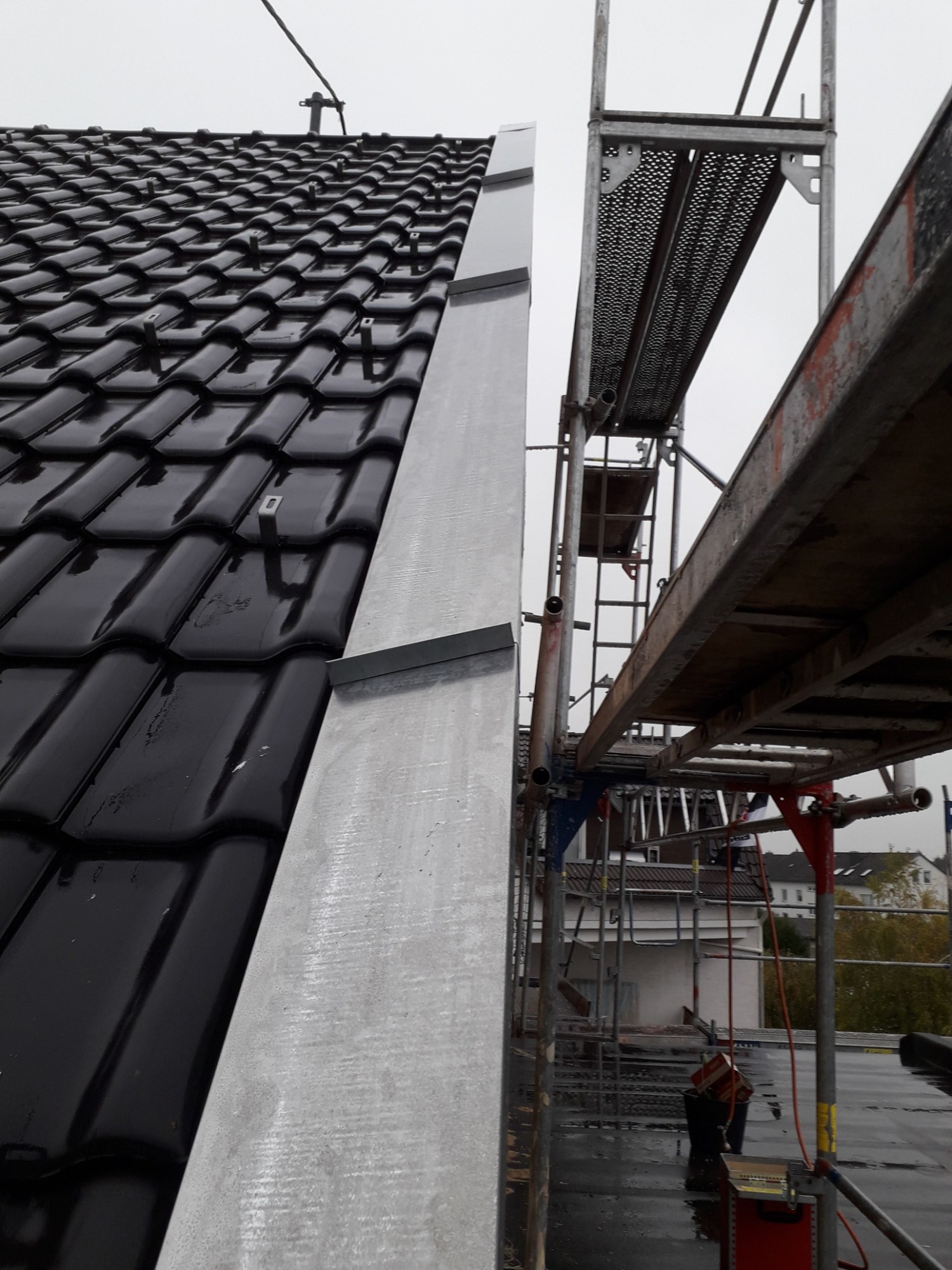 Błyszczący, czarny dach z dachówki ceramicznej, obróbka blacharska w kolorze srebrnym, rusztowanie w tle, pochmurny dzień.