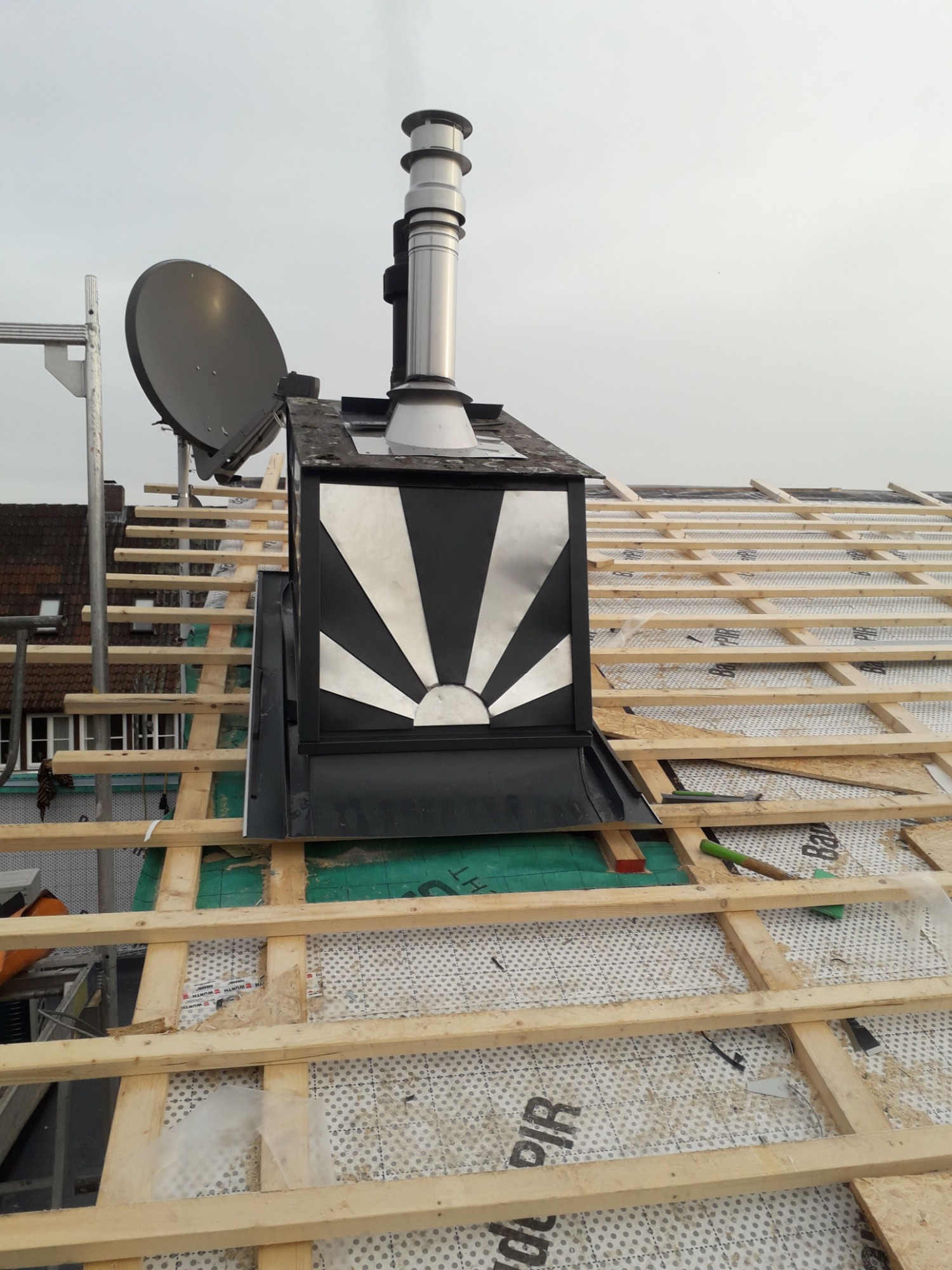 Konstrukcja dachu z drewnianymi łatami, widoczny komin z metalową obudową ozdobioną motywem słońca, antena satelitarna w tle.