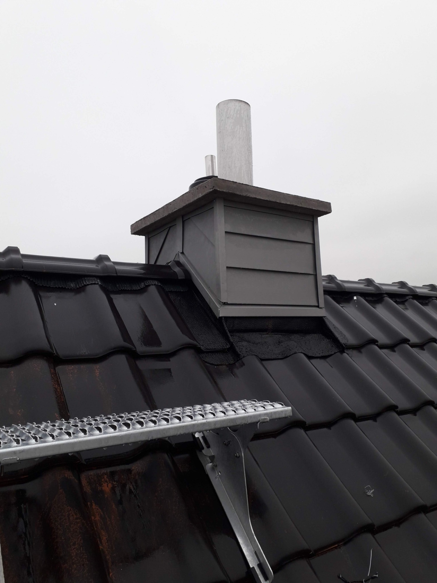 Błyszcząca, czarna dachówka ceramiczna z szarą obudową komina i fragmentem metalowej ławy kominiarskiej, widok z dołu pod kątem na tle pochmurnego nieba.