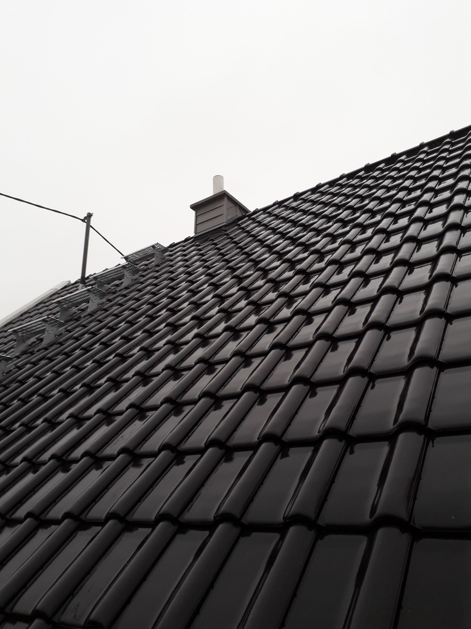 Błyszczący, czarny dach z ceramicznej dachówki z zamontowanymi stopniami kominiarskimi i kominem na tle pochmurnego nieba.