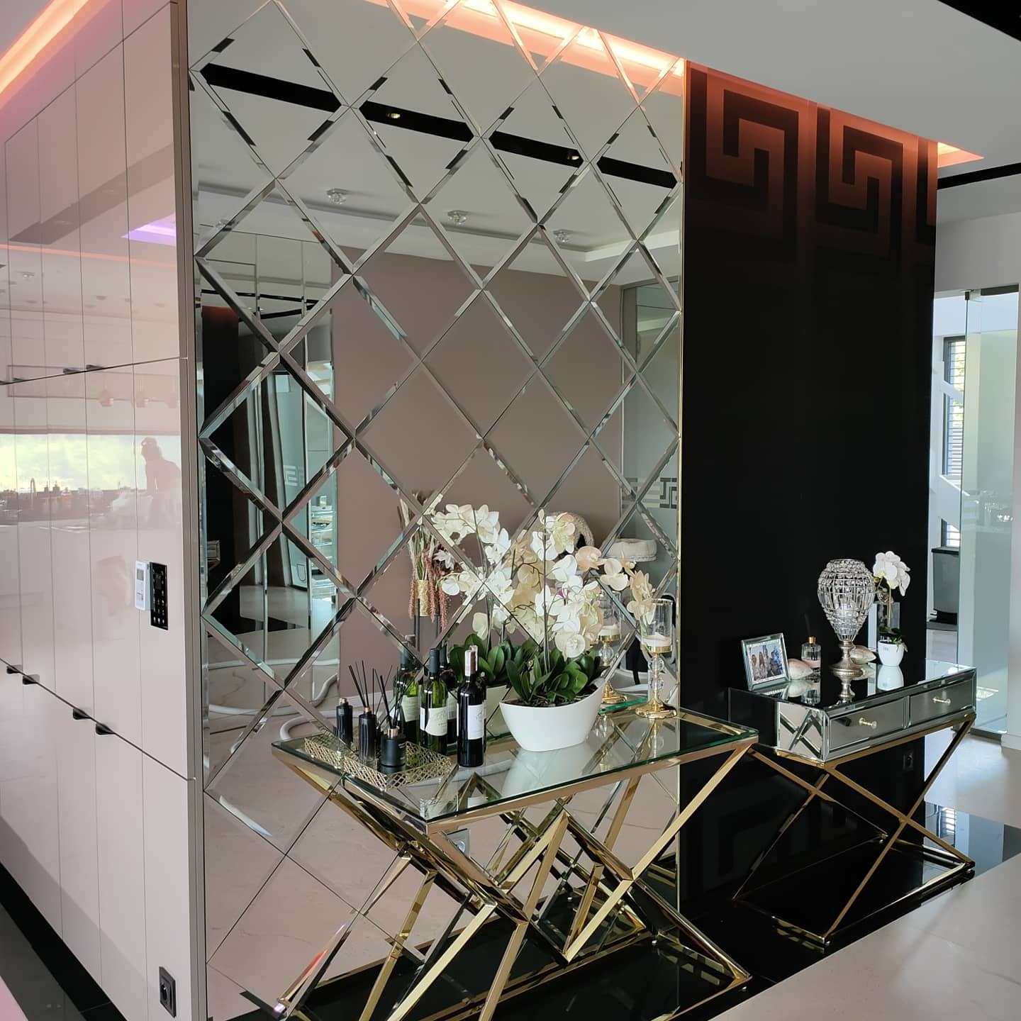 Ściana wyłożona lustrzanymi kwadratami z fazowanymi krawędziami, odbijająca wnętrze salonu z dekoracjami, ustawiona obok czarnej ściany z geometrycznym wzorem i jasnych szafek.