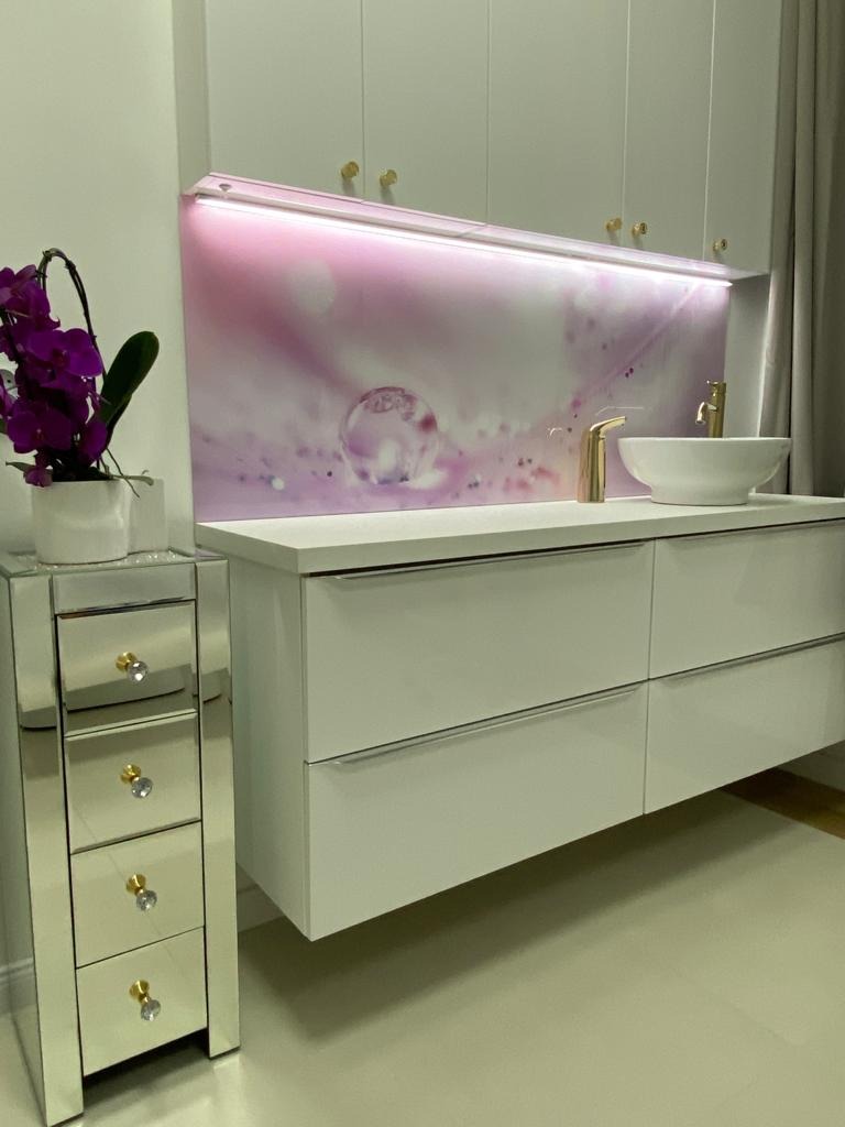 Biała, podwieszana szafka łazienkowa z umywalką nablatową i złotą armaturą, oświetlona panelem szklanym z motywem kropli wody w odcieniach różu, obok lustrzana komoda z kwiatami.