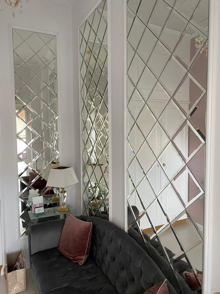 Eleganckie lustrzane panele ścienne z fasetowanymi rombami, odbijające fragment szarej sofy z aksamitną poduszką i lampę stołową, tworzące iluzję przestrzeni w jasnym wnętrzu.