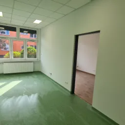 Pomalowane na jasnozielono wnętrze pokoju z oknem, przez które widać budynek i drzewa, oraz otwartymi drzwiami do kolejnego pomieszczenia z parkietem.