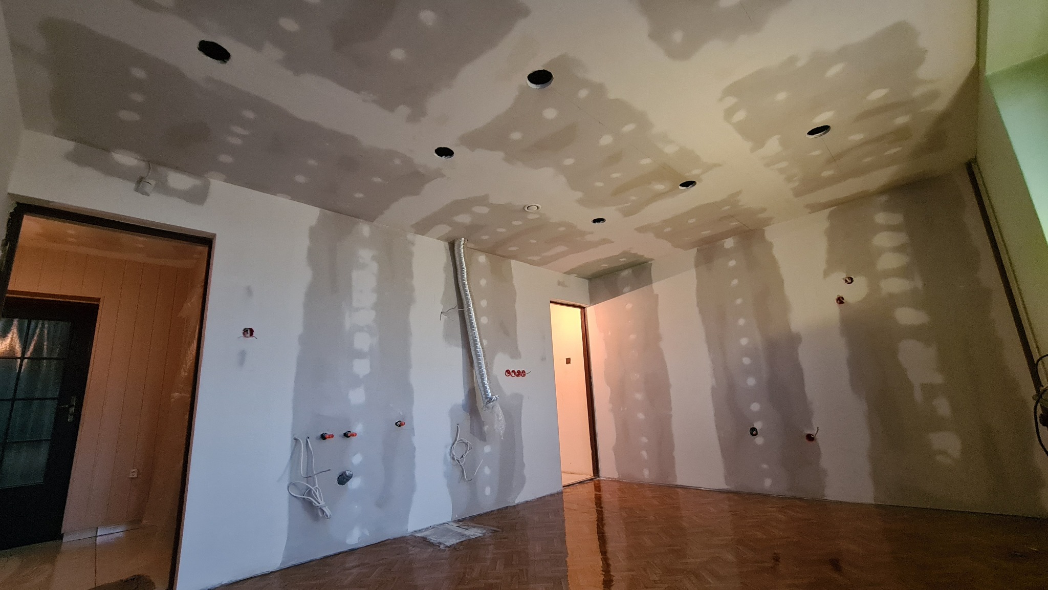 Pokój w trakcie remontu: ściany z nałożoną gładzią, widoczne otwory na oświetlenie w suficie, instalacja elektryczna na ścianach, parkiet na podłodze i rura wentylacyjna.