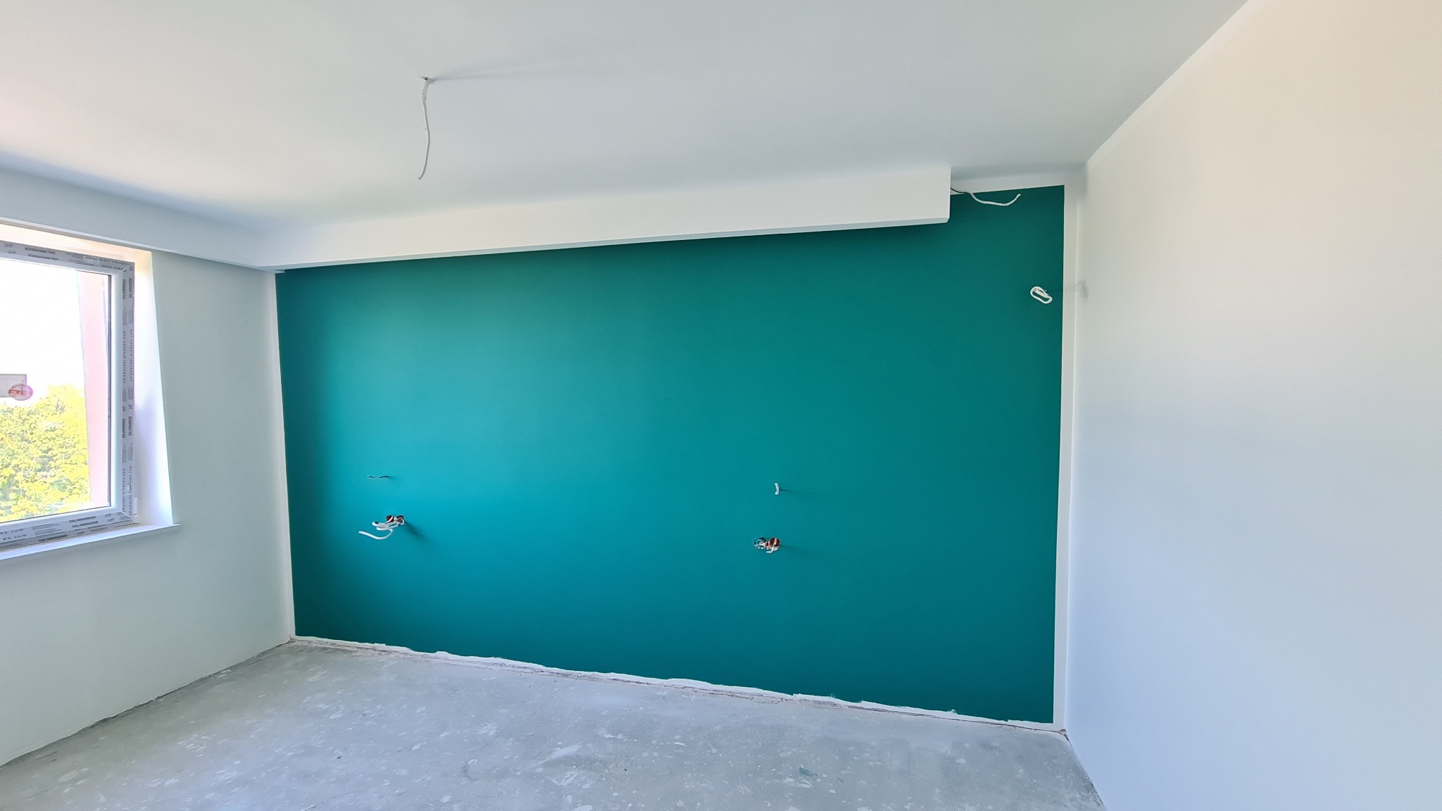Świeżo pomalowana na turkusowo ściana z widocznymi puszkami elektrycznymi, białe ściany boczne i sufit, surowa podłoga przed wykończeniem.
