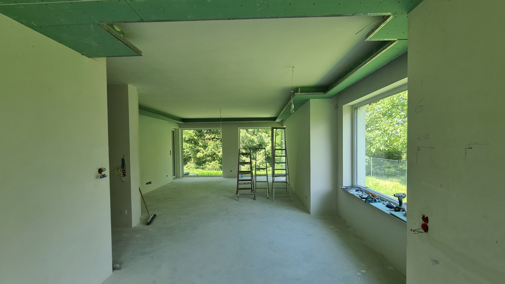 Wnętrze w trakcie remontu: surowy stan ścian z widocznymi instalacjami elektrycznymi, częściowo zamontowane płyty kartonowo-gipsowe na suficie, dwie drabiny ustawione w centralnej części...