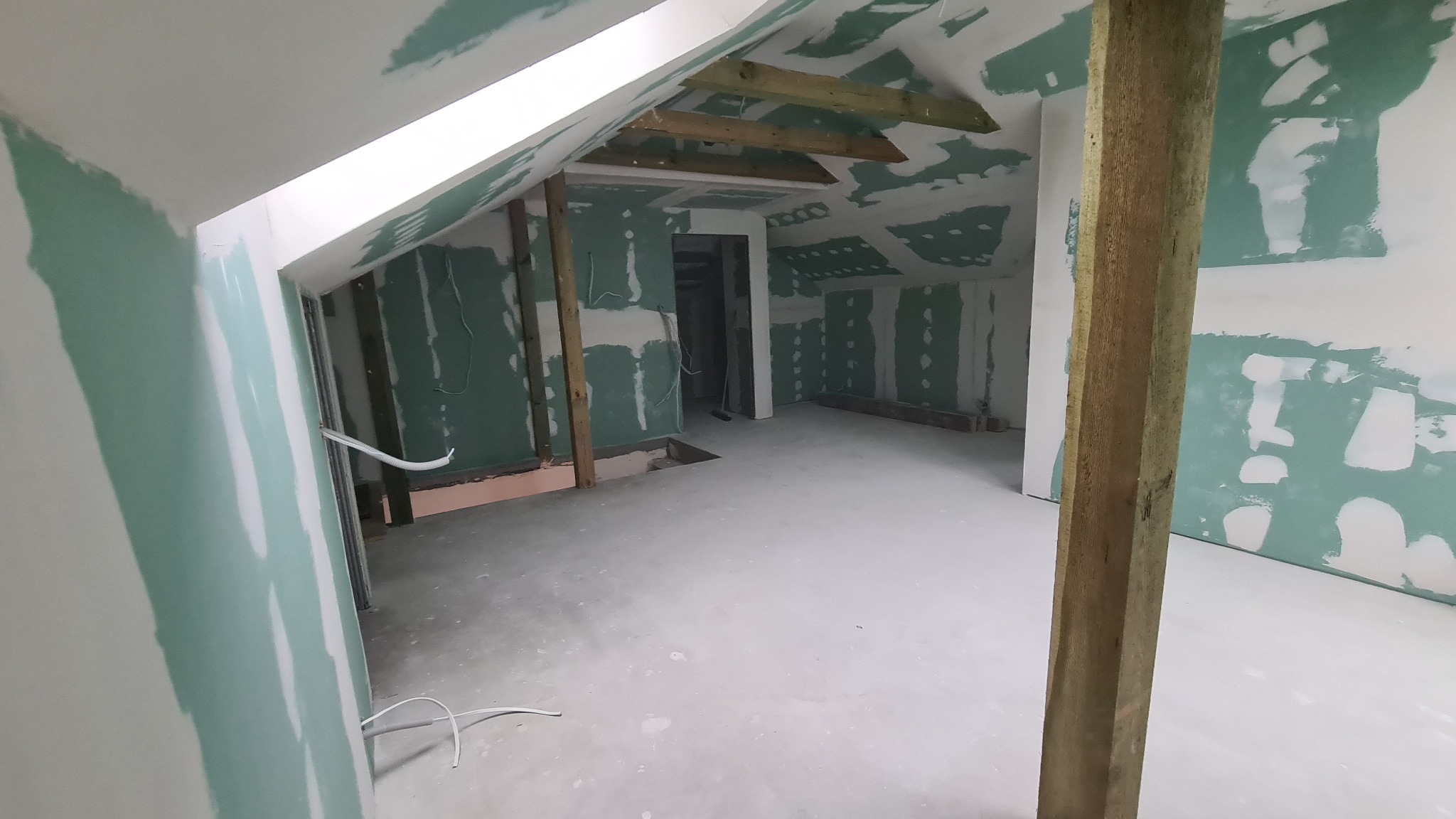 Poddasze w trakcie prac wykończeniowych: ściany pokryte płytami kartonowo-gipsowymi, widoczne drewniane elementy konstrukcyjne, przewody elektryczne wystające ze ścian, surowa wylewka podłogowa.
