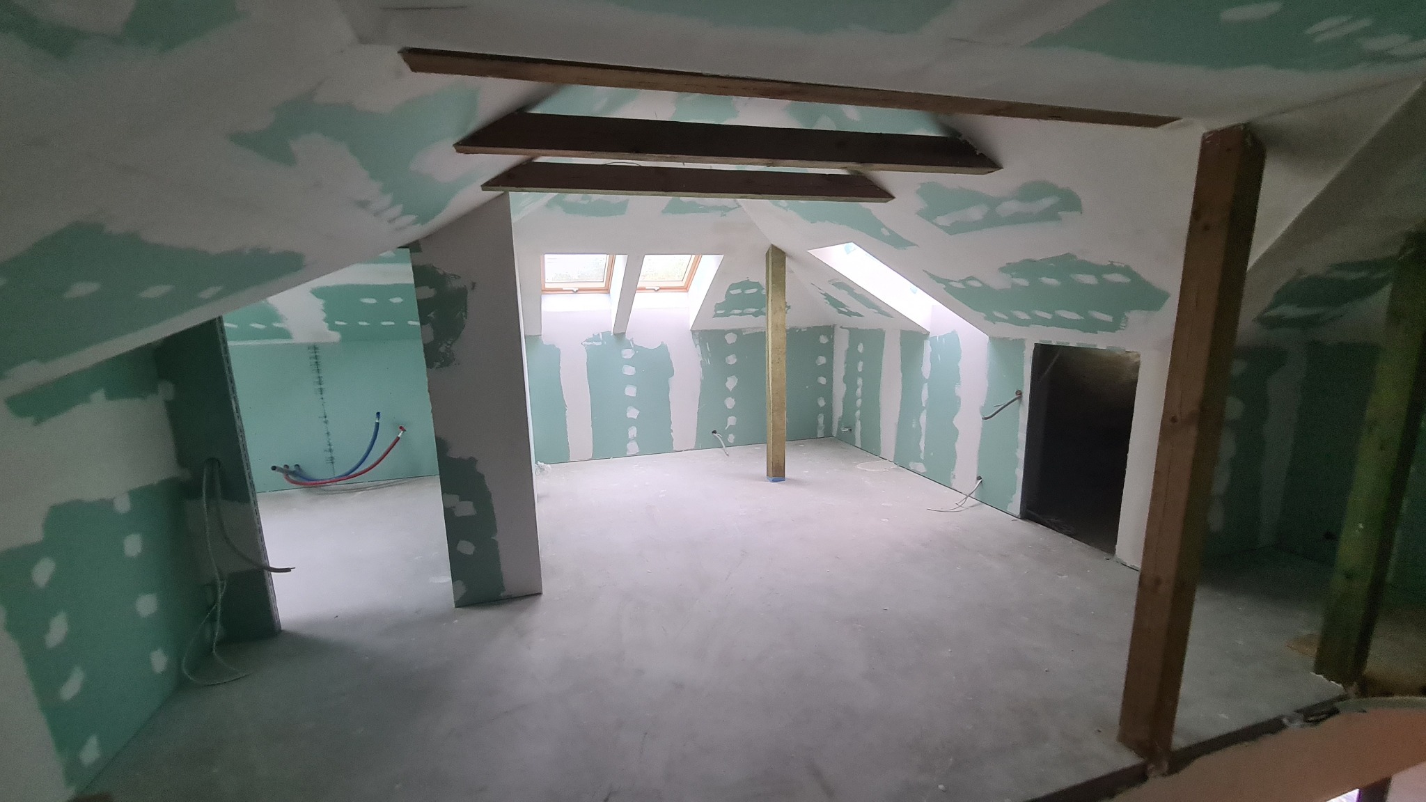 Poddasze w trakcie adaptacji: ściany wyłożone płytami kartonowo-gipsowymi, częściowo zaszpachlowane, widoczne drewniane elementy konstrukcyjne dachu i instalacje.