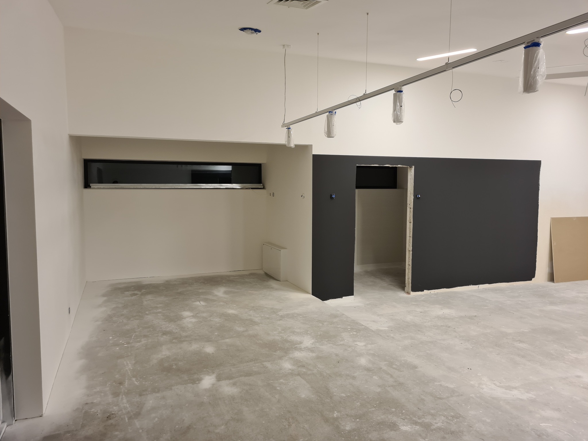 Wnętrze w trakcie remontu: surowe ściany pomalowane na biało i grafitowo, betonowa wylewka na podłodze, widoczne oprawy oświetleniowe zabezpieczone folią malarską, małe okno w ścianie.