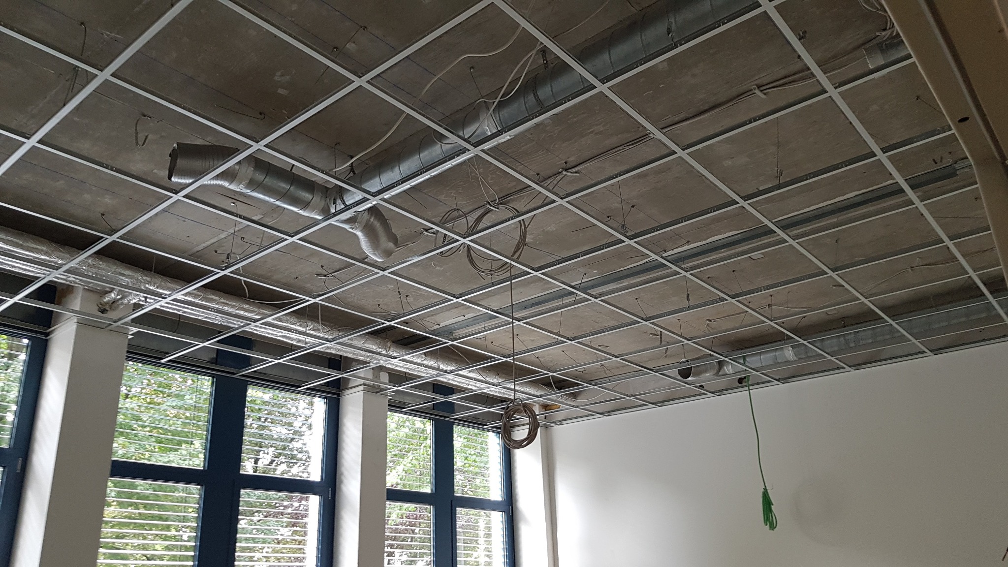 Sufit podwieszany w trakcie montażu, widoczne metalowe profile, przewody wentylacyjne i instalacje elektryczne. Duże okno z widokiem na zieleń w tle.