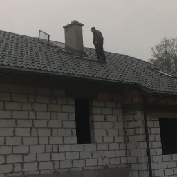 Dom jednorodzinny w trakcie krycia dachu z ciemnej dachówki, dekarz na dachu przy kominie, surowy stan elewacji z betonu komórkowego, pochmurna pogoda.