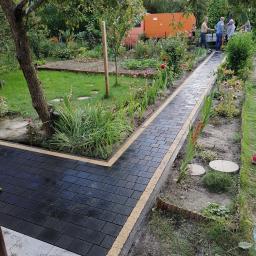 Profesjonaliści - Ścieżka z czarnej kostki brukowej z jasnymi obrzeżami w ogrodzie, widoczni ludzie w tle, częściowo ukończona realizacja.