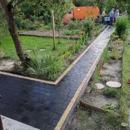 Ścieżka z czarnej kostki brukowej z jasnymi obrzeżami w ogrodzie, widoczni ludzie w tle, częściowo ukończona realizacja.