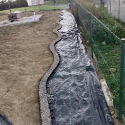 Profesjonaliści - Przygotowanie terenu pod budowę: czarna agrowłóknina rozłożona wzdłuż ogrodzenia, obrzeże z drobnego kamienia wyznaczające kształt przyszłej ścieżki, w tle sterta kostki brukowej.