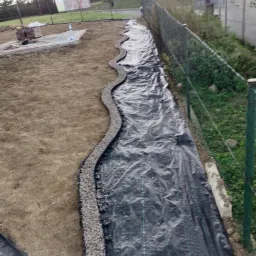 Przygotowanie terenu pod budowę: czarna agrowłóknina rozłożona wzdłuż ogrodzenia, obrzeże z drobnego kamienia wyznaczające kształt przyszłej ścieżki, w tle sterta kostki brukowej.