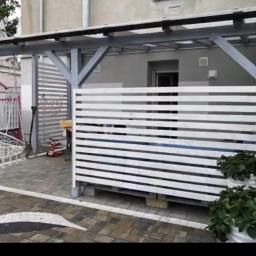 Profesjonaliści - Nowoczesny taras z drewnianą pergolą i białymi panelami poziomymi, wykończony kostką brukową z dekoracyjną białą linią, donice z roślinami na tle budynku.