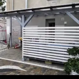 Nowoczesny taras z drewnianą pergolą i białymi panelami poziomymi, wykończony kostką brukową z dekoracyjną białą linią, donice z roślinami na tle budynku.