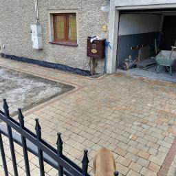 Profesjonaliści - Nowo ułożona kostka brukowa przed domem, z widocznym fragmentem czarnego ogrodzenia i głową psa w kadrze, prowadząca do otwartego garażu z taczką i narzędziami w środku.