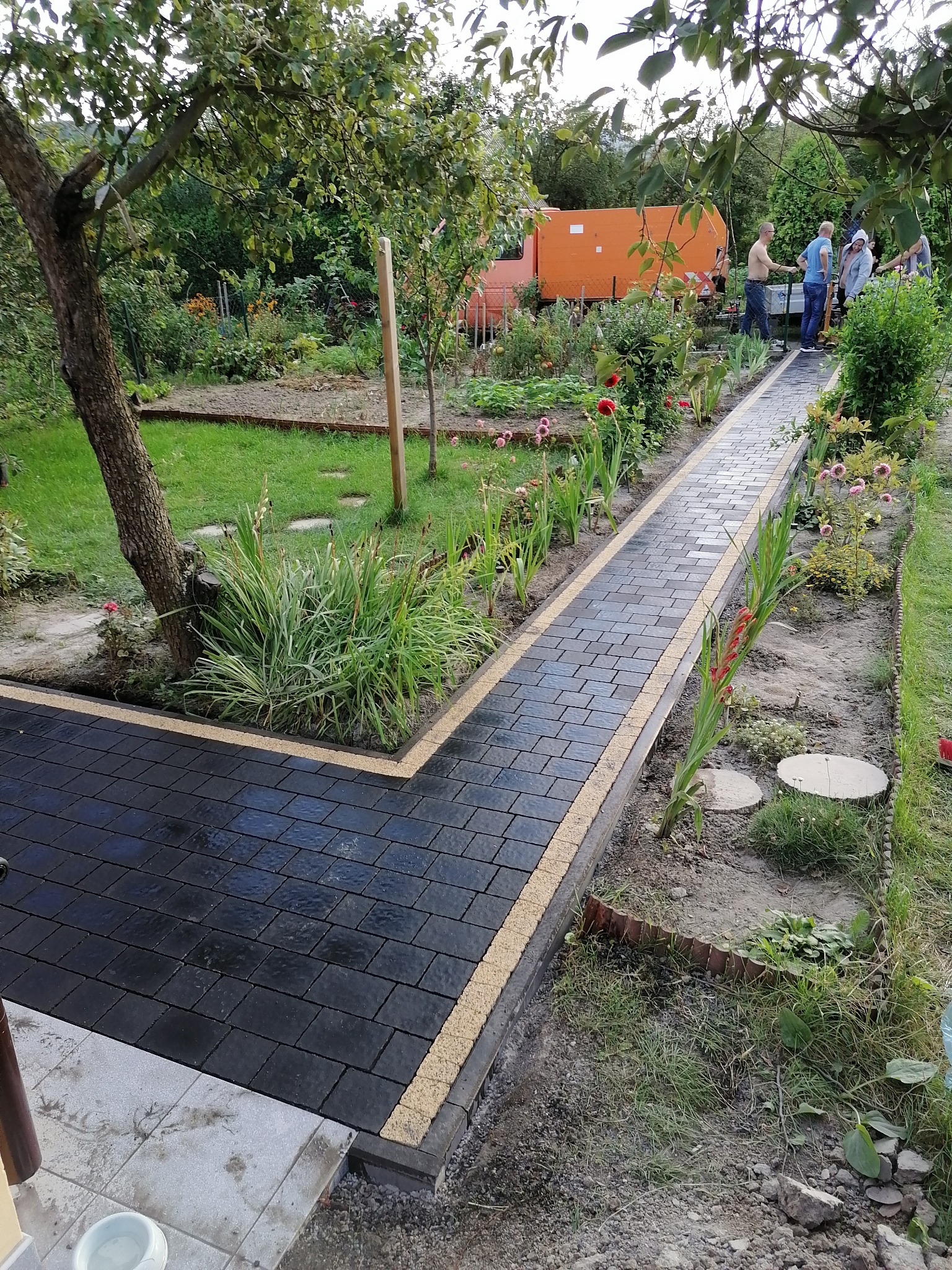 Ścieżka z czarnej kostki brukowej z jasnymi obrzeżami w ogrodzie, widoczni ludzie w tle, częściowo ukończona realizacja.