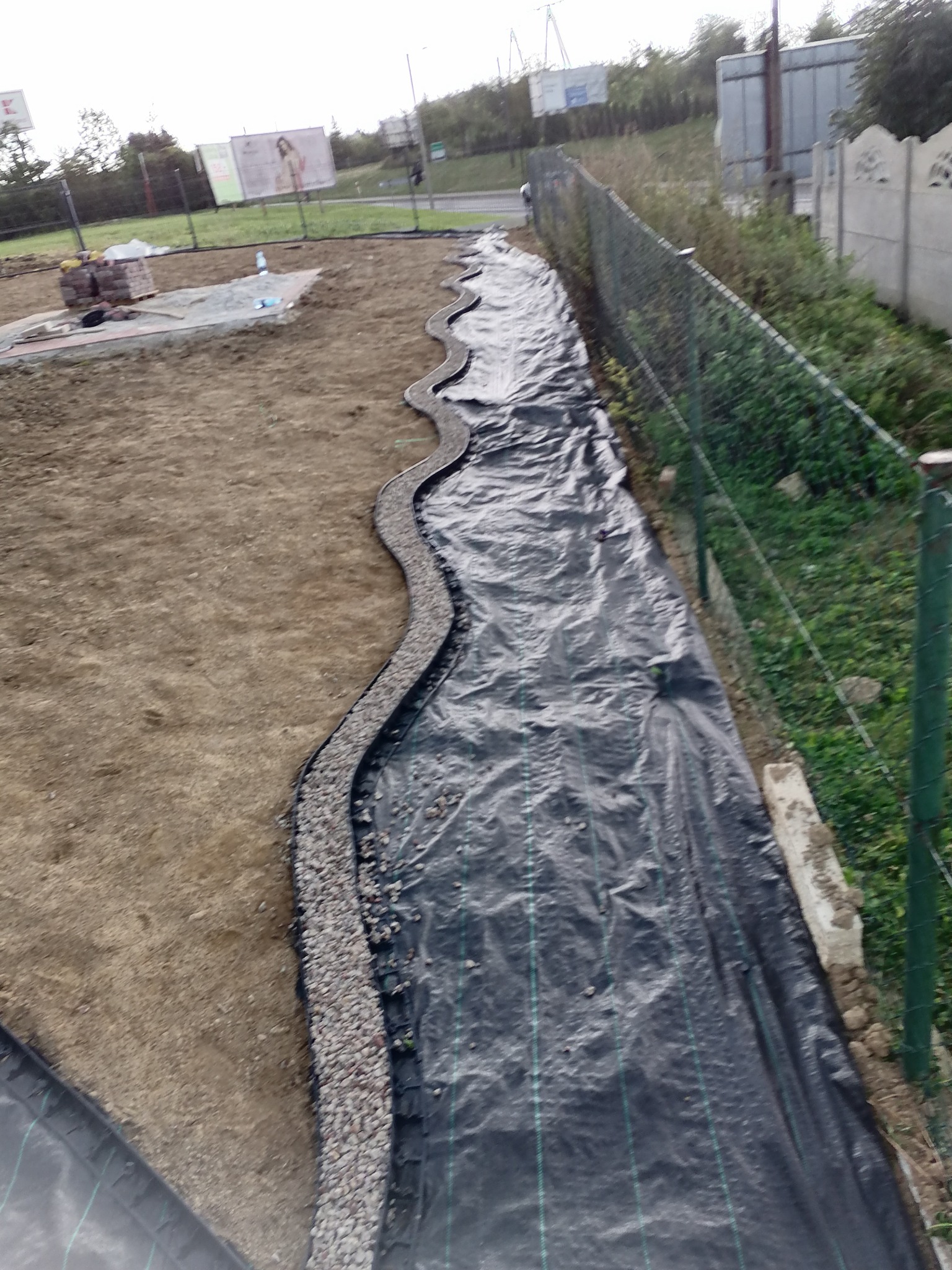 Przygotowanie terenu pod budowę: czarna agrowłóknina rozłożona wzdłuż ogrodzenia, obrzeże z drobnego kamienia wyznaczające kształt przyszłej ścieżki, w tle sterta kostki brukowej.