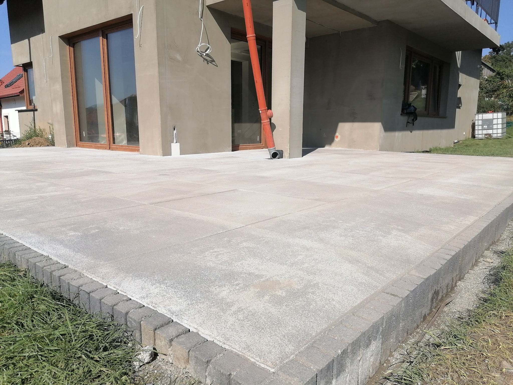 Wykończony taras z szarych płyt betonowych obrzeżony kostką przy domu w stanie surowym zamkniętym, widoczna rynna spustowa i fragment trawnika.